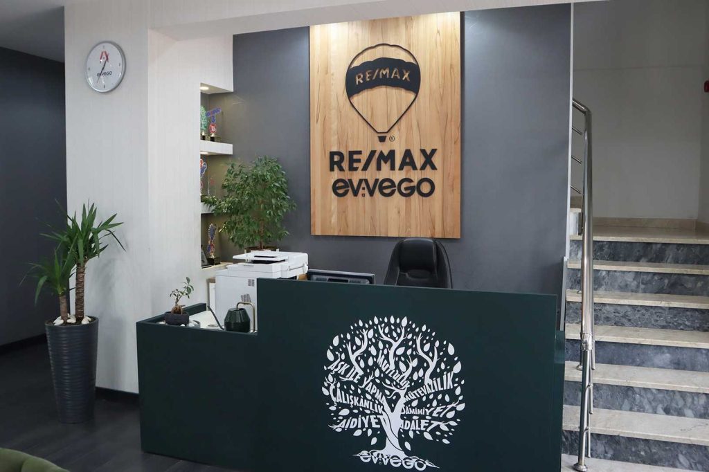 RE/MAX Evvego ofisinin modern ve şık resepsiyon alanı. Ahşap duvarda büyük RE/MAX logosu ve ofisin marka kimliğini yansıtan detaylar bulunuyor.